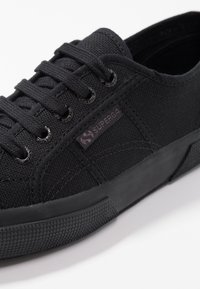 Baskets en toile noires avec un dessus texturé, une semelle en caoutchouc et des lacets ronds, arborant une petite étiquette logo sur le côté.