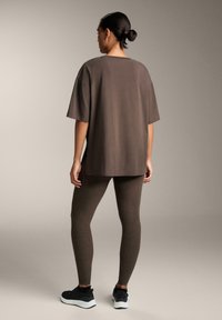 Barna, nagym�éretű póló rövid ujjakkal, együttesen illeszkedő barna leggings-el és fekete sportcipőkkel, hátulról nézve.