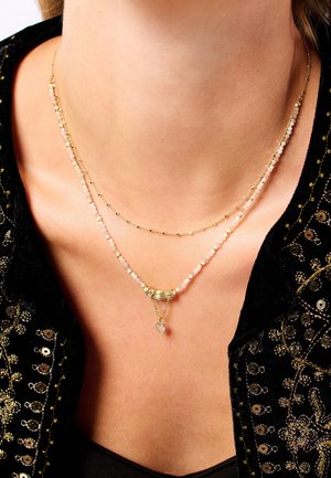 Collier superposé plaqué or avec de petites perles opalescentes, un élément central métallique et un pendentif en forme de cœur. Détails de surface texturée visibles.