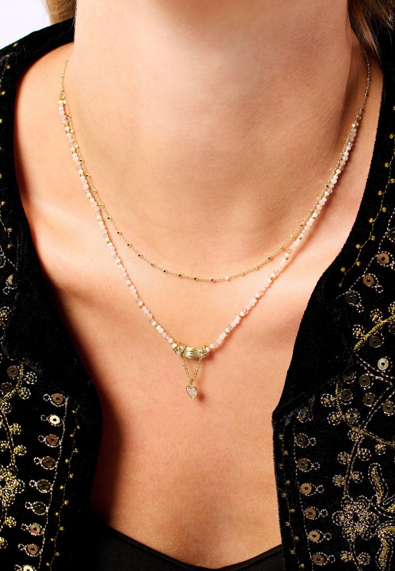 Collier superposé plaqué or avec de petites perles opalescentes, un élément central métallique et un pendentif en forme de cœur. Détails de surface texturée visibles.