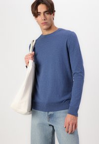 Blue sweater in soft fabric, crew neck, and ribbed cuffs, paired with light denim jeans and a beige tote bag. Simple, streamlined design.
