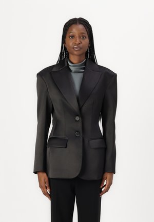 Sort satin blazer med strukturerede skuldre, to frontlommer og to knapper. Båret over en mørk, højhalset top, parret med sorte bukser.