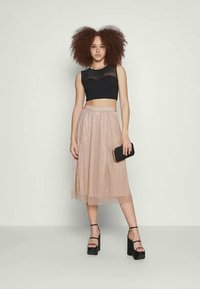 Top preto cropped com detalhes transparentes, combinado com uma saia plissada até ao joelho de tom rosa pálido com uma textura brilhante. Modelo segura uma pequena carteira preta.