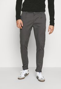 Pantalones de algodón gris tipo chino con un corte slim, que presentan un cierre de botón en la parte frontal y puños enrollados, combinados con zapatillas blancas y una camiseta negra de manga larga.