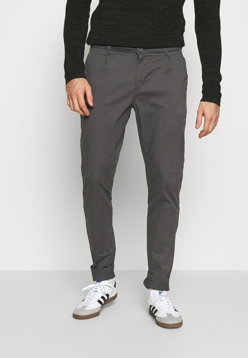 Pantalones de algodón gris tipo chino con un corte slim, que presentan un cierre de botón en la parte frontal y puños enrollados, combinados con zapatillas blancas y una camiseta negra de manga larga.