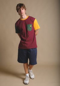 T-shirt z wielokolorowymi blokami w odcieniach burgundowym, zielonym i żółtym, z przednią kieszenią z logo słonia. W połączeniu z granatowymi szortami i trampkami.