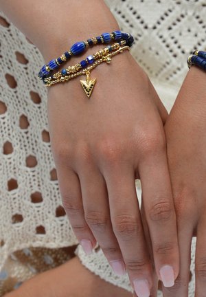 Bunte Armband-Set mit tiefblauen Glasperlen, goldenen Akzenten und einem goldenen dreieckigen Charm auf einem Handgelenk vor einem Spitzenhintergrund.