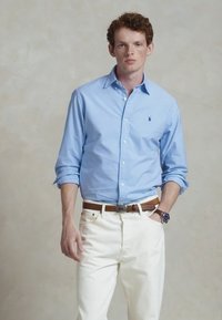 Šviesiai mėlynas marškinėlių viršus su ilgomis rankovėmis, derinamas su baltais kelnėmis. Aprangą papildo rudas diržas ir laikrodis.