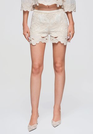 Kvinde iført beige blonde shorts og matchende spidse hæle står foran en ensfarvet baggrund.