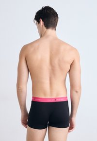 Boxer shorts noirs avec une ceinture "TOMMY HILFIGER" rose. Tissu lisse, design ajusté, détails minimaux. Personne de dos par rapport à la caméra.