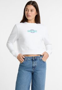 Valkoinen lyhythihainen kangaspusero, jossa on monivärinen "Guess Jeans" -logo edessä. Yhdistetty korkeavyötäröisiin sinisiin farkkuihin, joissa on rento leikkaus.