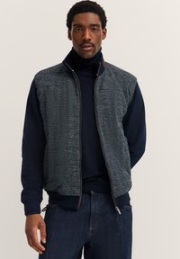 Uomo che indossa un maglione a collo alto blu navy, un gilet grigio a spina di pesce con zip e jeans blu scuro su uno sfondo semplice chiaro.