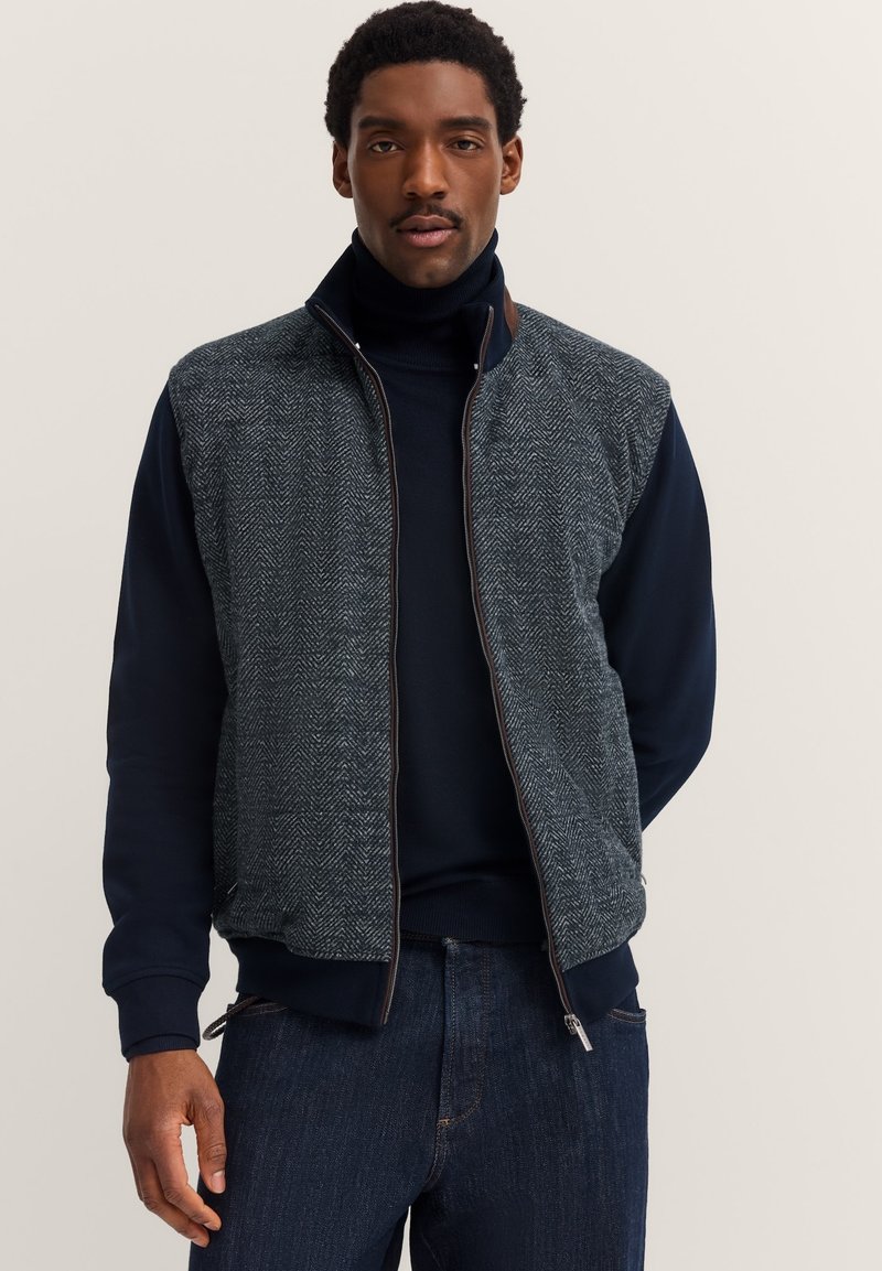 Uomo che indossa un maglione a collo alto blu navy, un gilet grigio a spina di pesce con zip e jeans blu scuro su uno sfondo semplice chiaro.