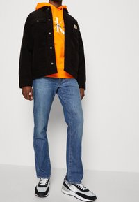 Svart kordjakke med trykknapper lagd over en oransje hettegenser. Lys blå straight-leg jeans og svart-hvite sneakers fullfører looken.