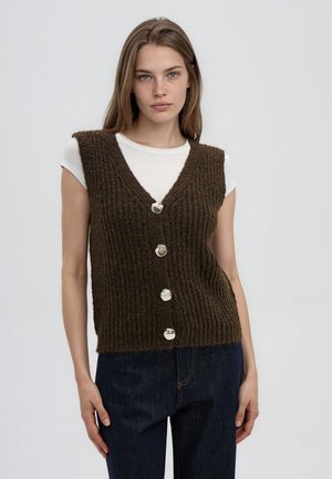 Giovane donna che indossa una camicia bianca a maniche corte sotto un gilet marrone lavorato a maglia con bottoni e jeans blu scuro su uno sfondo semplice.