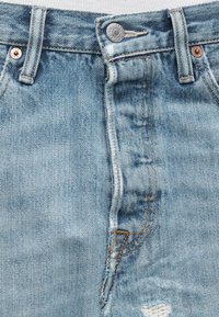 Κοντινό πλάνο σε ανοιχτό μπλε τζιν παντελόνι με κουμπωτή μέση, θηλιές για ζώνη και εμφανή φθαρμένα ράμματα κοντά στο φερμουάρ.