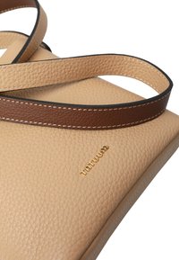 Bež usnjena torba z teksturirano površino, opremljena s temno rjavim trakom in zlatim logotipom. Dizajn je pravokoten z nežnimi šivi kot poudarki.