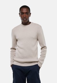 Pull en tricot côtelé beige avec un col rond et un détail de quatre boutons sur l'épaule. Il dispose de manches longues et de poignets ajustés.