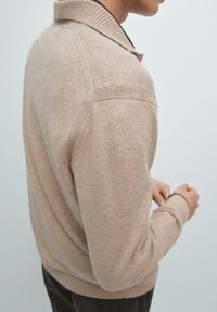 Maglione beige lavorato a maglia con scollo a collo, polsini a coste e finitura testurizzata; presenta lievi variazioni di colore e una silhouette aderente.