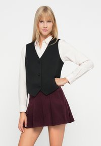Blonde vrouw in een witte button-up shirt, zwarte knopen vest en een geplisseerde donkerpaarse rok, poseert met één hand op de heup.