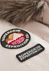 Brūns audums ar ūdens pilieniem; satur divus izšūtus uzlīmes. Viens attēlo "Superdry Everest" ar kalna dizainu, bet otrs "Superdry Expedition Co."