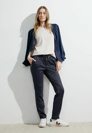 Maglione beige chiaro, blazer blu navy e pantaloni jogger grigio scuro con coulisse. Presenta sneakers bianche con accenti rossi e verdi. Texture liscia.