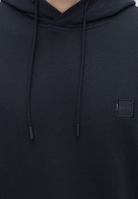 Mørk marineblå hættetrøje med snore og et lille firkantet "BOSS" logo-patch på brystet, close-up af overkroppen.