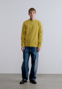 Κίτρινο φούτερ με ριμπ μανσέτες και τελείωμα, συνδυασμένο με φαρδιά μπλε τζιν. Το μοντέλο στέκεται σε γκρι δάπεδο μπροστά σε ανοιχτόχρωμο φόντο.