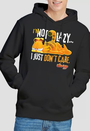 Ein junger Mann lächelt und trägt einen schwarzen Hoodie, der eine gezeichnete faule Katze mit dem Text "ICH BIN NICHT FAUL... ICH INTERESSIERE MICH EINFACH NICHT." zeigt.