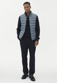 Pelēks pūkains veste ar stāvu apkakli, uzvilkta virs melnas džemperes. Saskaņota ar tumšiem biksēm un melniem apaviem, izstādīta pret baltu fona.