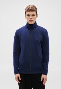 Tumši zils sporta krekls ar rāvējslēdzēju, augstu apkakli un ribotām aprocēm. Uz krūtīm neliela logotipa uzlīme, izgatavots no mīksta, teksturēta auduma.