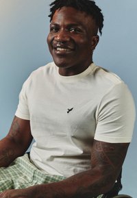 Fehér, rövid ujjú póló, fekete madárlogóval a mellrészen, zöld kockás rövidnadrággal párosítva. Az egyén karján látható tetoválások vannak.