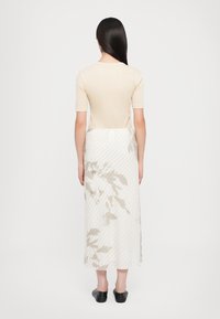 Beige eng anliegendes Oberteil mit kurzen Ärmeln, kombiniert mit einem mittellangen, weißen Rock mit abstrakten Blumenmustern und diagonalen Streifen. Schwarze Schuhe.