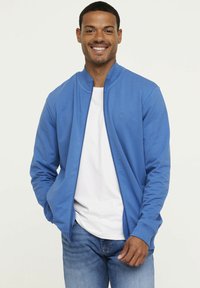Veste zippée bleue en tissu lisse, avec un col rond, des poignets côtelés et un logo sur la poitrine. Associée à un t-shirt blanc en dessous.