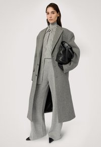 Šedý oversized kabát s texturovaným povrchem, spárovaný se světle šedým rolákem a pruhovanou košilí, šedými kalhotami s širokými nohavicemi a černou kabelkou.