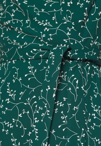 Grüner Stoff mit einem weißen Blumenmuster. Verfügt über ein gebundenes Gürtel-Detail, eine glatte Textur und ein fließendes Design.