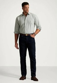 Mężczyzna stoi, ubrany w jasnoszary, zapinany na guziki, koszulę z podwiniętymi rękawami, ciemne dżinsy, brązowy pasek, brązowe buty i zegarek na nadgarstku, patrząc w prawo.