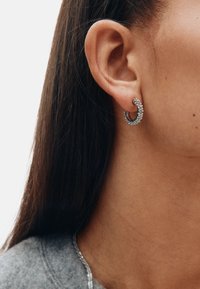 Srebrne naušnice u obliku obruča s prozirnim, sjajnim detaljima prikazane su nose na ušnoj školjci, uz ravnu tamnu kosu i sivu majicu.