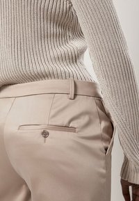 Beige Hose mit einer glatten Textur, ausgestattet mit einer Gesäßtasche mit Knopf, kombiniert mit einem hellgrauen Rippenpullover.