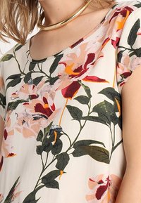 Donna che indossa una blusa crema a fantasia floreale con grandi fiori rosa, rossi e arancioni e foglie verdi, abbinata a una collana a cerchio dorata.
