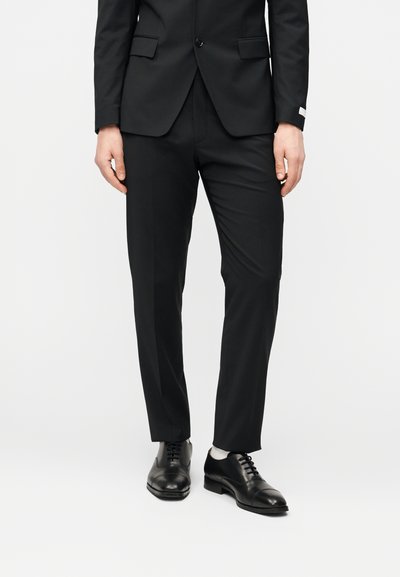 Ανδρικό μαύρο κοστούμι με παντελόνι στενής γραμμής, σε συνδυασμό με γυαλιστερά μαύρα δερμάτινα παπούτσια. Διαθέτει καθαρές γραμμές και ομαλή υφή υφάσματος.