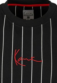 Tricou negru cu dungi verticale albe, având un guler rotund cu nervuri și un logo brodat roșu pe față. Mărimea L etichetată în interior.