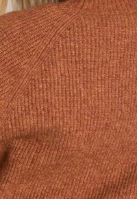Gros plan sur une personne portant un pull en tricot texturé de couleur rouille avec des motifs de côtes visibles et des coutures diagonales.