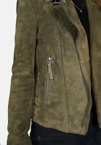 Olive green suede jacket featuring a zip closure, side zipper pockets, and a textured finish. Has an asymmetrical hem and metallic accents.