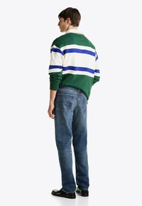 Ung mand står i profil iført en grøn, hvid og blå stribet rugbytrøje, blå jeans og sorte loafers på en hvid baggrund.