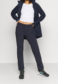 Marine waterdichte jas over een witte cropped t-shirt, gecombineerd met donkere broeken en zwarte sportschoenen met groene accenten.