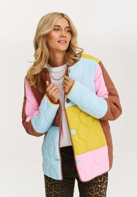 Žena s dlhými blond vlasmi, oblečená v viacfarebnej prešívanej bunde a nohaviciach s leopardím vzorom, upravuje si golier bundy a pozerá sa do strany.