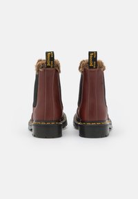 Botas de cuero marrón hasta el tobillo con laterales elásticos negros, ribete de pelo en el cuello y costura amarilla en la suela de goma negra.