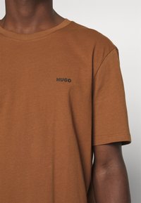 Bruine T-shirt met korte mouwen en een klein zwart "HUGO" logo op de linkerborst, gedragen door een persoon met een donkere huid.