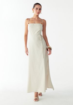 Lange lichtbeige jurk met een vierkante halslijn, verstelbare bandjes, een zijsplit en een tailledetail met strik. Gecombineerd met minimalistische sandalen.